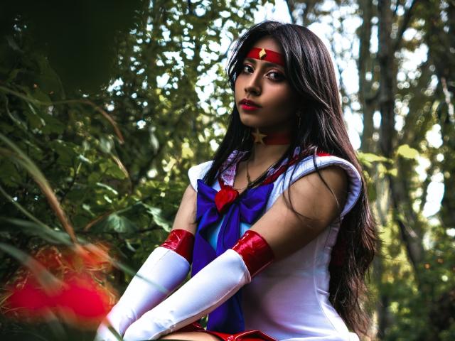 VioletXBloom wears a Sailor Moon cosplay, with a white sleeveless top, red and blue bow, and white gloves with red accents. She has long dark hair, red lipstick, and a red headband with a gold star. Her expression is neutral, with slightly parted red lips. The forest background is blurred, focusing on VioletXBloom. The lighting is natural, highlighting her facial features and costume. The image is sharp, with vivid colors and a realistic style.