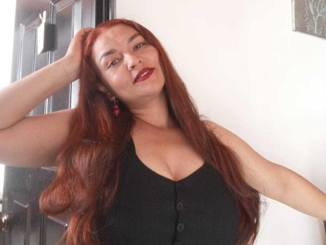 VivianBig tiene el pelo largo y rojo, lleva una blusa negra abotonada que deja ver sus pechos, aretes rojos y lápiz labial rojo, y está sonriendo con una mano en el pelo..