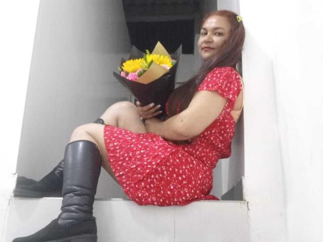 VivianBig, con cabello rojo largo, vestido rojo, botas negras, sosteniendo un ramo de flores, sentada en un marco de ventana blanco, sonriendo, sosteniendo el ramo contra su trasero, piernas dobladas, mirando a la cámara..