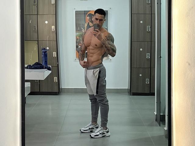 Uxiox sin camisa, con tatuajes, pantalones grises, zapatillas blancas, sosteniendo un teléfono, de pie en un vestuario, selfie frente al espejo, mostrando abdominales marcados, brazos musculosos, cabello negro corto, expresión seria..