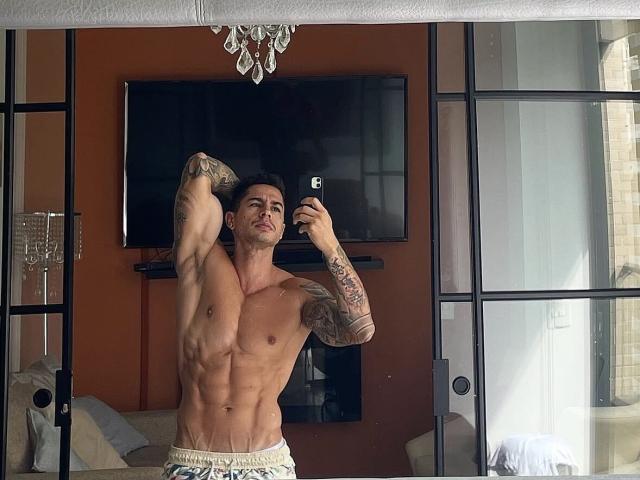 Uxiox, sin camisa, musculoso, tatuado, flexionando el bíceps, sosteniendo un teléfono, selfie en el espejo, pantalones cortos blancos, abdominales marcados, piel clara, cabello corto y oscuro, expresión seria, de pie en una habitación luminosa..