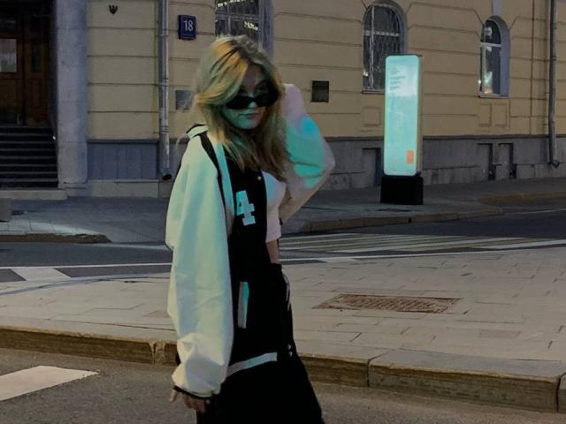 ChristopherSzala, a blonde woman with wavy hair, wears a black and white varsity jacket and dark pants. She has a black mask covering her nose and mouth. She walks on a city street at night, holding a mask to her face. The street has a lighted sign and a beige building with arched windows in the background. The lighting is dim, with a slight greenish-blue glow. The composition is straightforward, focusing on ChristopherSzala in the foreground.