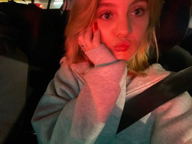 Close-up of ChristopherSzala in a car. Blonde hair, fair skin, red lipstick, and light makeup. Wearing a grey hoodie, left hand on cheek, red nail polish. Seatbelt across chest. Warm red and yellow light on face. Soft texture of hoodie. Right eye slightly open, left eye closed. Ring on left hand. Slightly blurred background. Casual, intimate selfie.