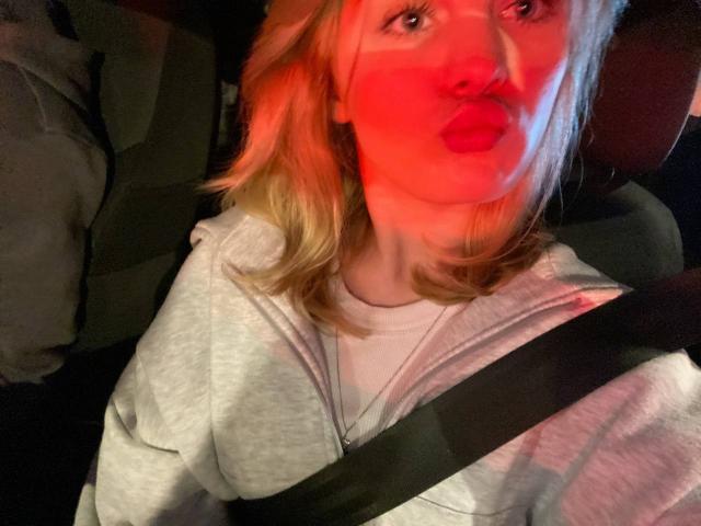 Close-up photo of ChristopherSzala with blonde hair, wearing a grey hoodie, and a seatbelt across the chest. Red and blue lighting casts dramatic shadows on ChristopherSzala's face, emphasizing a pouty expression with slightly parted lips. The image has a dynamic, almost surreal quality due to the contrasting lights. ChristopherSzala's gaze is directed upwards, adding a sense of curiosity or tension. Textures include the soft, slightly fuzzy fabric of the hoodie and the smooth, slightly reflective surface of the seatbelt.