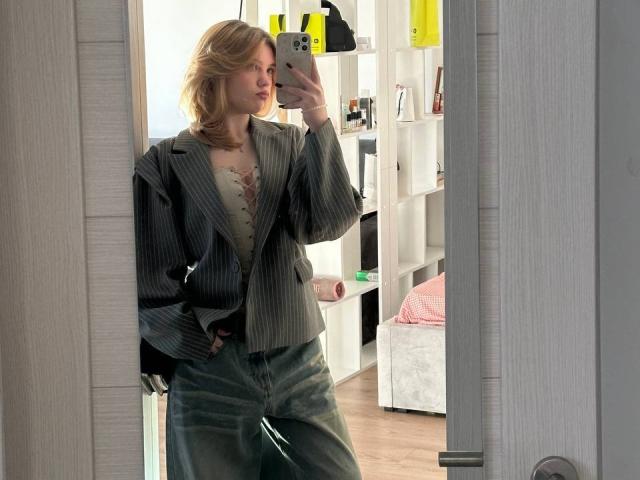 ChristopherSzala stands in a mirror, taking a selfie. He wears a pinstriped blazer over a lace-up, beige top and loose, greenish-gray pants. His blonde hair is wavy, and he holds a silver phone in his right hand. The reflection shows his left hand in his pocket. The room has white shelves with yellow and black objects and a pink bed. The mirror is framed by a gray, wooden door.