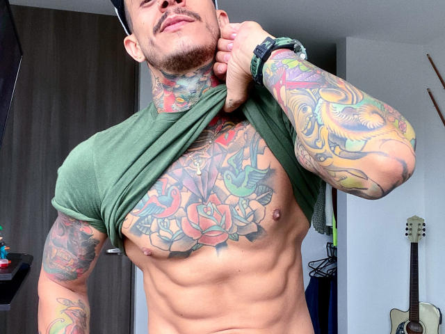 FitNolan with colorful tattoos, lifting green shirt to reveal chiseled abs and chest tattoos, wearing black watch, in a room with a guitar.