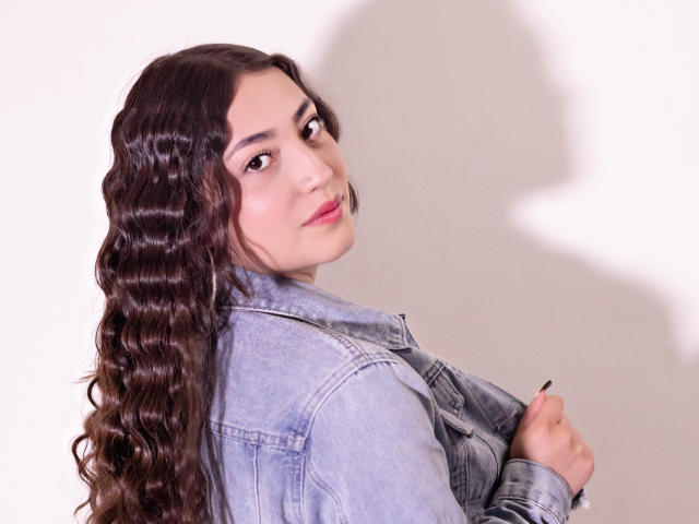 AnnFisher with wavy brown hair, looking over shoulder, wearing a blue denim jacket, holding it with one hand, shadow on white wall, slightly smiling, pink lipstick.