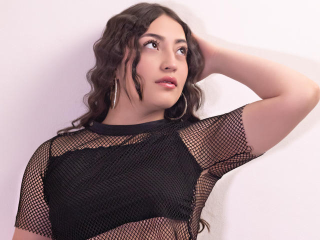 AnnFisher with wavy brown hair, wearing a black mesh top, looking to the side, hand in hair, hoop earrings, soft pink lipstick, slight smile, visible small tits.
