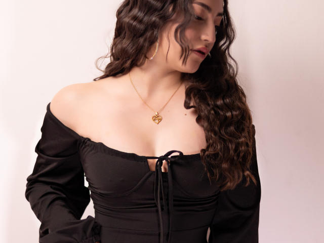 AnnFisher with wavy brown hair, wearing a black off-shoulder dress, gold necklace with a knot pendant, and showing her tits. She's looking down, arms at her sides.