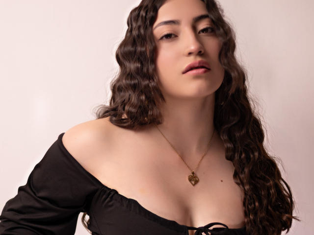 AnnFisher with wavy brown hair, wearing a black off-shoulder top, showing her tits, and a gold heart necklace, has a neutral expression, and is looking slightly to the side.