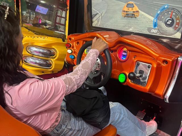 MyaaTyler sits in a bright orange toy truck's driver's seat, holding the steering wheel with her right hand. She wears a pink, long-sleeve, lace-trimmed top and blue jeans. The dashboard is orange with colorful buttons and a green light. A digital speedometer shows 96. The truck's exterior is yellow with dual headlights. The image focuses on MyaaTyler's back and the truck's interior. The setting is indoors, likely a toy store.