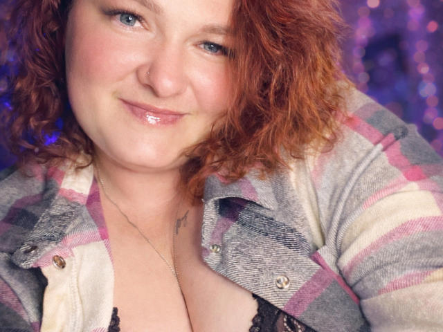 AngelQSky with curly red hair, pinkish cheeks, and a smile, wearing a plaid shirt, exposing her large, shiny tits in a black lace bra, against a purple background.