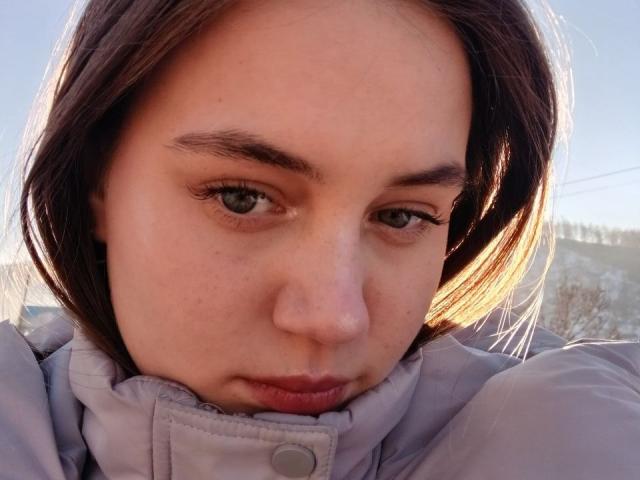 AlysiaFinkelstein heeft een lichte huid, bruine ogen en kort bruin haar, en draagt een lichtgrijze jas met een knoopsluiting. Ze kijkt naar beneden met een licht fronsende uitdrukking op een foto met veel zonlicht..