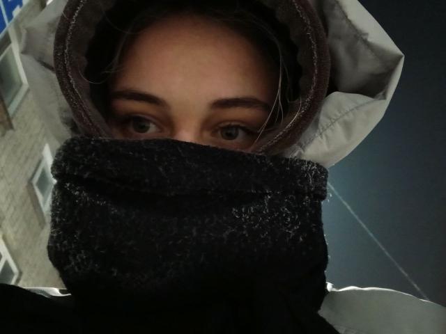 Het gezicht van AlysiaFinkelstein, met donkere ogen, een bruine kap en een zwart masker dat de helft van haar gezicht bedekt, een nachtelijke setting, ze kijkt omhoog, met een licht nerveuze uitdrukking, een subtiele gloed van een straatlamp..