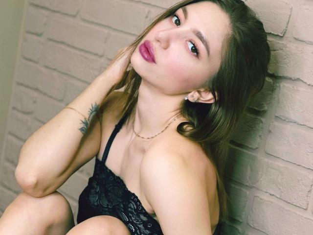 GreatKat with long brown hair, red lipstick, and black lace lingerie, sitting against a white brick wall, hand on neck, showing a tattoo on her upper arm, looking sexy.