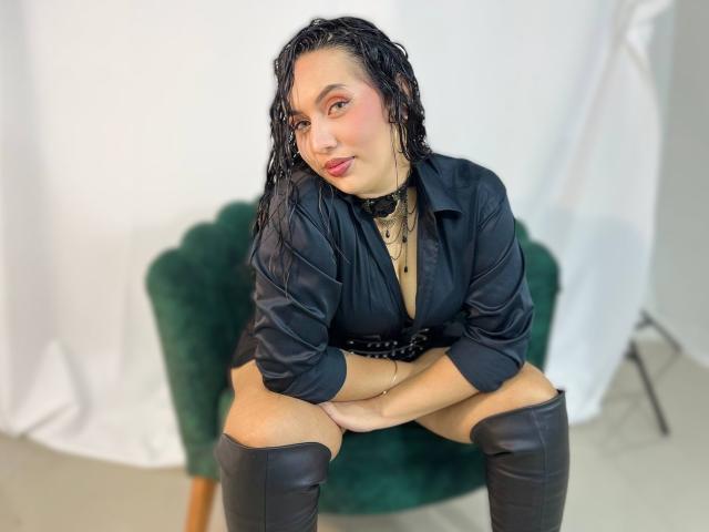 AmyRoss with wet black hair, red eyeshadow, black shirt, choker, black boots, sitting on green chair, legs apart, hands on knees, looking at camera, smiling.