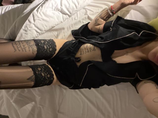 SensualMila lies on a bed with white sheets, wearing a black satin robe with white trim, partially open to reveal black lace stockings and black lace panties. Her tattooed arms and midsection are visible, with text tattoos on her left thigh and side. She has tattoos on her right forearm. Her right hand, adorned with a red bracelet, rests above her. The image focuses on her lower body and lingerie, emphasizing the texture of the lace and satin.