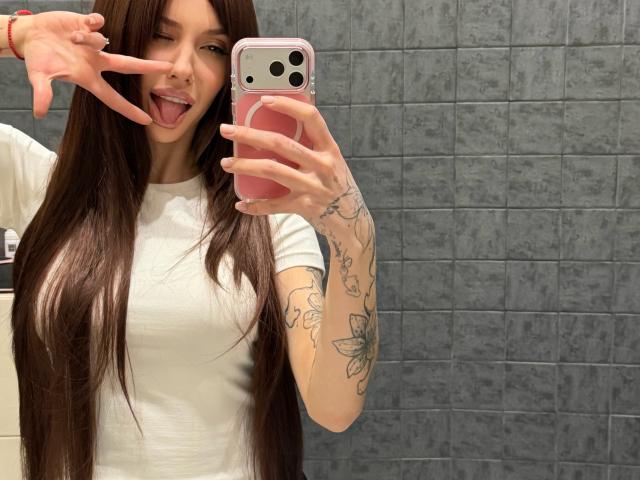 SensualMila takes a selfie in a bathroom, her long brown hair flowing over her shoulders. She sticks out her tongue and makes a playful "V" with her right hand. She holds a pink phone with a white camera lens in her left hand. She's wearing a white t-shirt that fits snugly, highlighting her small to medium breasts. Her tattooed arms are visible, featuring floral designs. The image is sharply focused with a gray tiled wall in the background. SensualMila's expression is playful and confident.