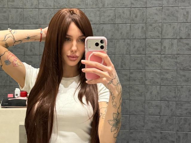 SensualMila, a young woman with long, straight brown hair, takes a mirror selfie. She holds a pink phone case in her right hand, her left arm raised with tattoos visible on both arms, including a butterfly and floral designs. She wears a white t-shirt, her lips slightly parted. Her skin is fair, and she has a nose ring. The background is a gray tiled wall, with a white counter and red and white bottles on it. The composition focuses on her reflection and her tattoos.