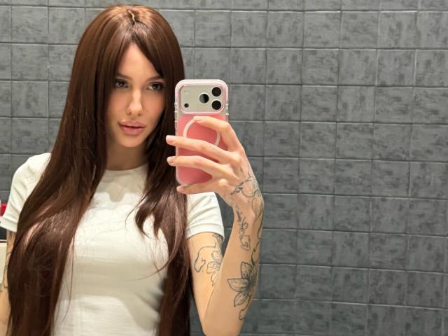 SensualMila, a young woman with long brown hair, stands in front of a mirror taking a selfie. She wears a white T-shirt, revealing tattoos on her right arm and left wrist. Her pink phone case with a white and pink design contrasts with her fair skin. The gray tiled background is slightly textured. Her expression is neutral, lips slightly parted. Her tattoos include floral and geometric patterns. The image focuses on her upper body and face.