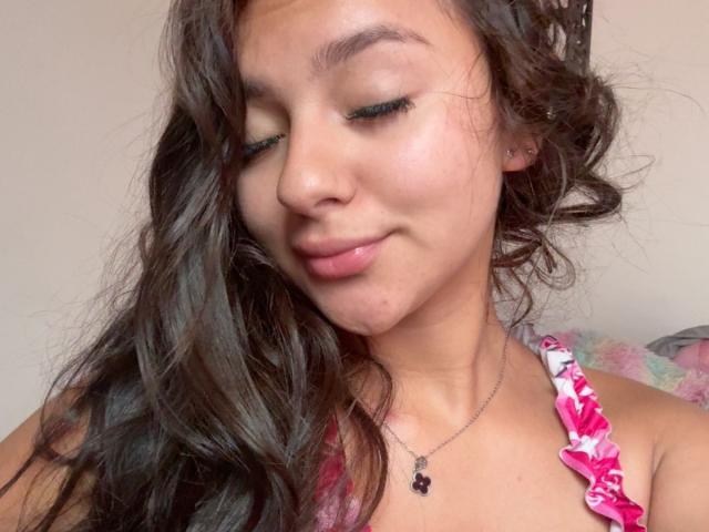 AlessiaFerry, con lunghi capelli castani ondulati, eyeliner verde e una maglietta rosa con fiori, sorride leggermente, mostrando una piccola imperfezione sulla guancia, e indossa una collana di perline nere..