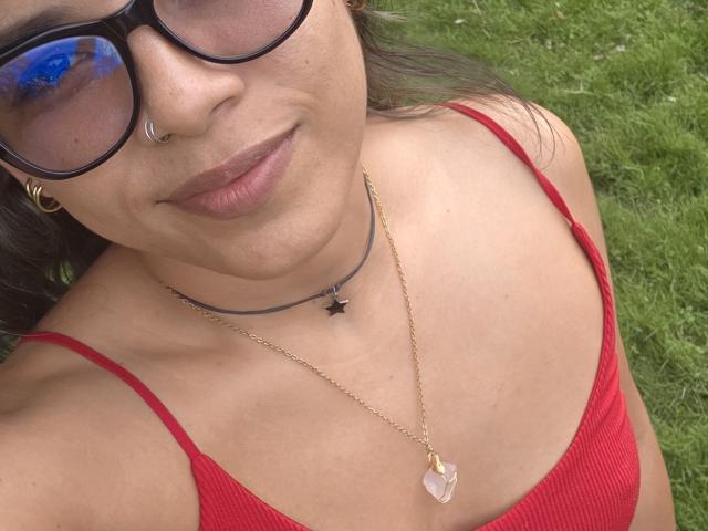 Close-up of LauraSan69. She has light brown skin, wears black-framed glasses, and has a nose ring. She's in a red ribbed tank top with thin straps, showing her collarbone. She wears a gold chain necklace with a small black cross and a larger pink crystal pendant. Her lips are slightly parted with a neutral pink lipstick. The grass in the background is green and slightly blurred. The image is focused on her upper body and face. Her dark hair is partially visible.