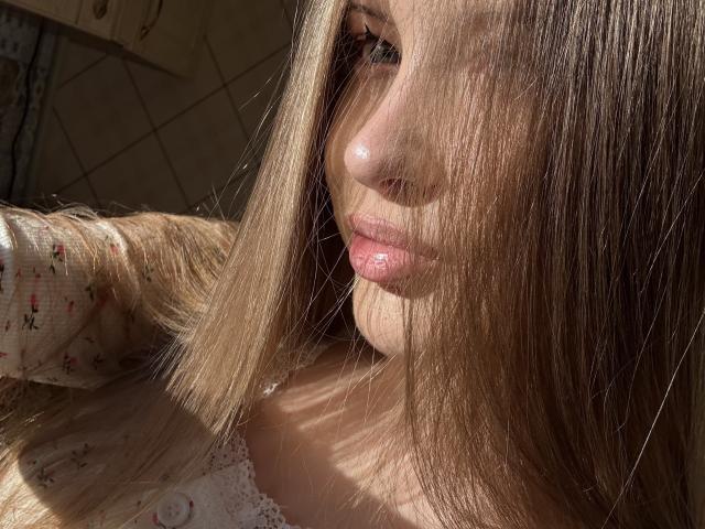 JoelleCanal's face with sunlit blonde hair, shiny pink lips, and a slight smile, wearing a floral top, in a bright, sun-drenched setting.