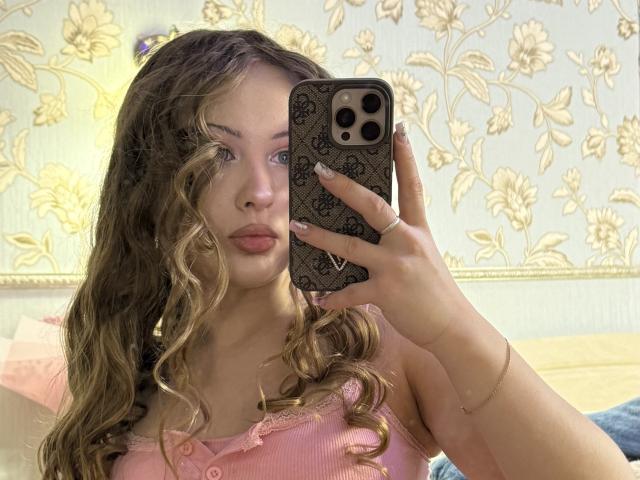 JoelleCanal with curly blonde hair, pink ribbed top, holding iPhone with black floral case, taking selfie, showing soft pink lips and subtle makeup. Boobs partially visible.