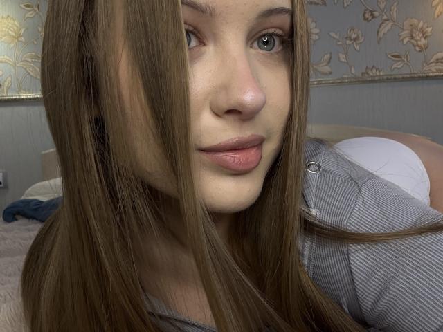 JoelleCanal with long brown hair, blue eyes, and pink lips, wearing a striped shirt, lying on a bed in white panties, showing her ass.