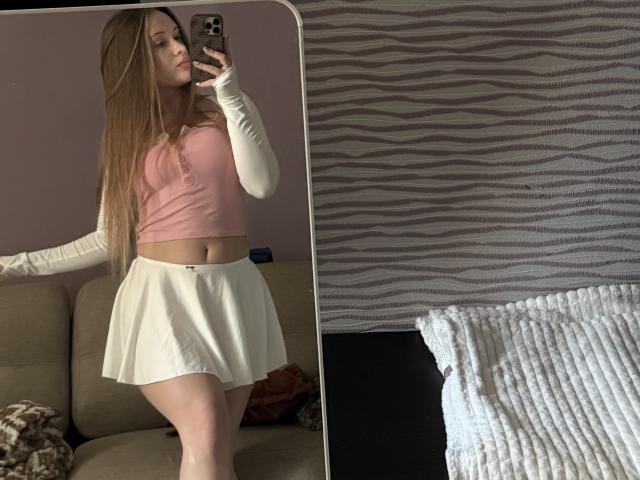 JoelleCanal with long blonde hair, pink crop top, white skirt, and white sleeves, taking a mirror selfie, showing her ass and midriff, in a casual, home setting.