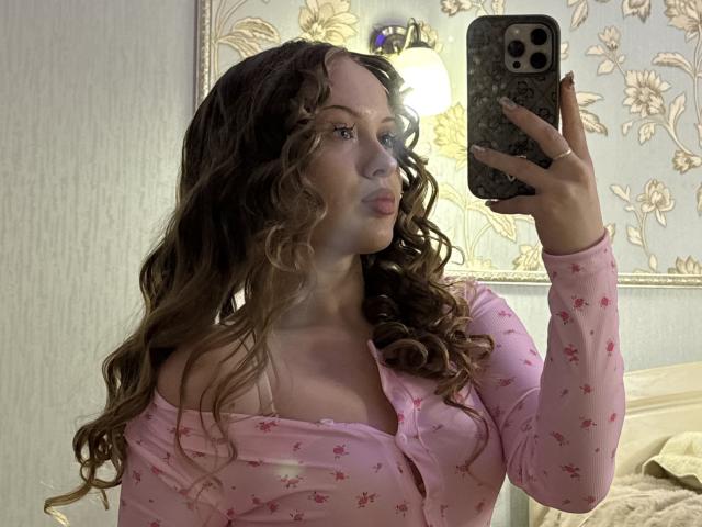 JoelleCanal with wavy brown hair, pouty lips, wearing pink floral long-sleeve top, taking mirror selfie with iPhone, showing her tits in the reflection.