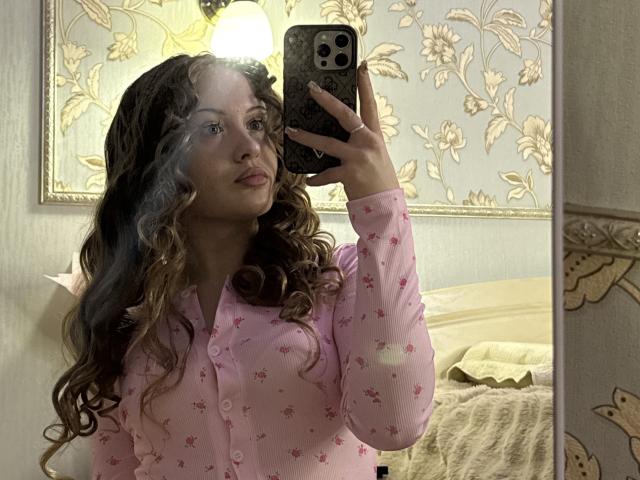 JoelleCanal with wavy brown hair, wearing pink floral pajamas, taking a mirror selfie with an iPhone, showing her face and upper body, in a dimly lit room.