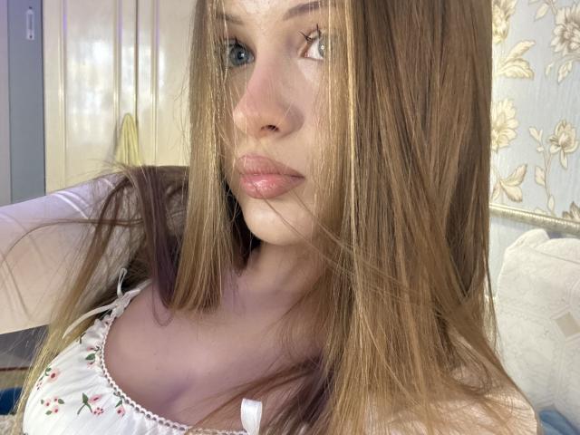 JoelleCanal has long, straight brown hair, blue eyes, and pink lips, wearing a white floral top, with sunlight filtering through her hair. Her tits are partially visible.
