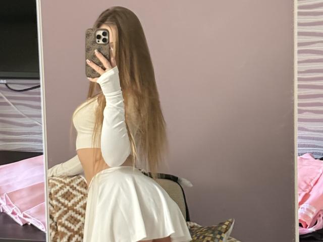 JoelleCanal with long brown hair, wearing a white crop top and long sleeves, white skirt, taking a mirror selfie, showing her ass, holding a phone with a patterned case.