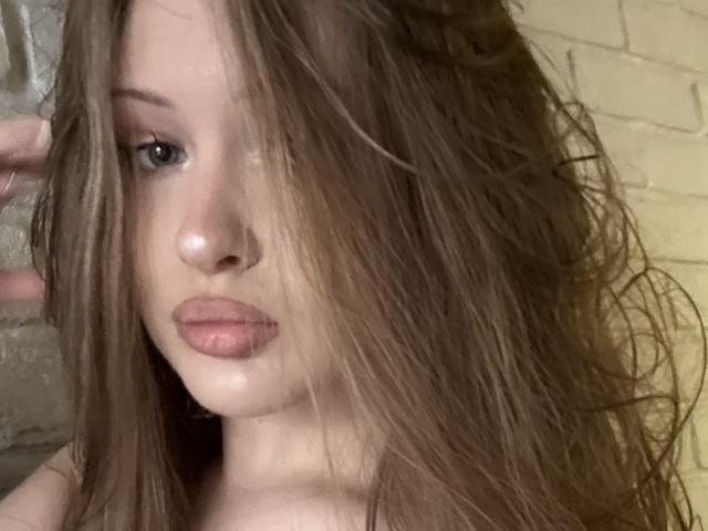 JoelleCanal has wet brown hair covering one eye, pale skin, and slightly parted pinkish lips. Her blue eyes are visible. Her hand is partially in the frame.