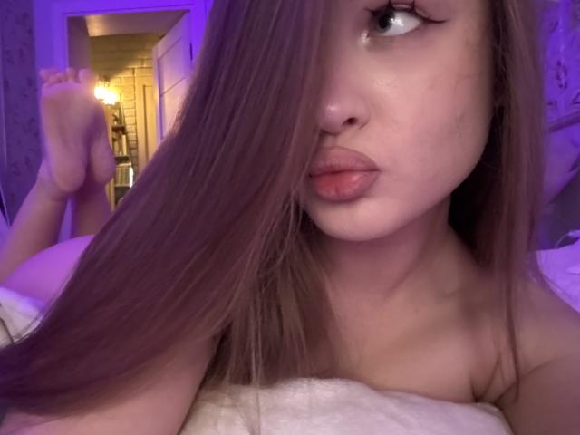 JoelleCanal with long brown hair covering her right eye, pouty lips, and bare shoulders, feet up in the background, purple-lit room, soft white blanket.