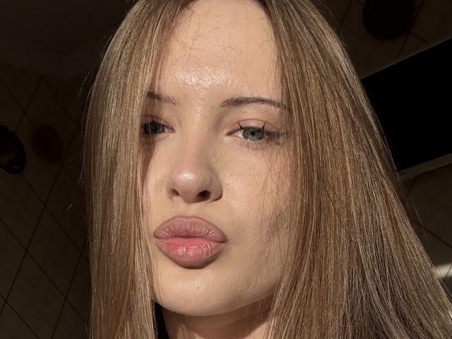 JoelleCanal's face, sunlit with shiny skin, straight brown hair, and pink pouty lips, showing a subtle pout, with green eyes looking slightly to the side.