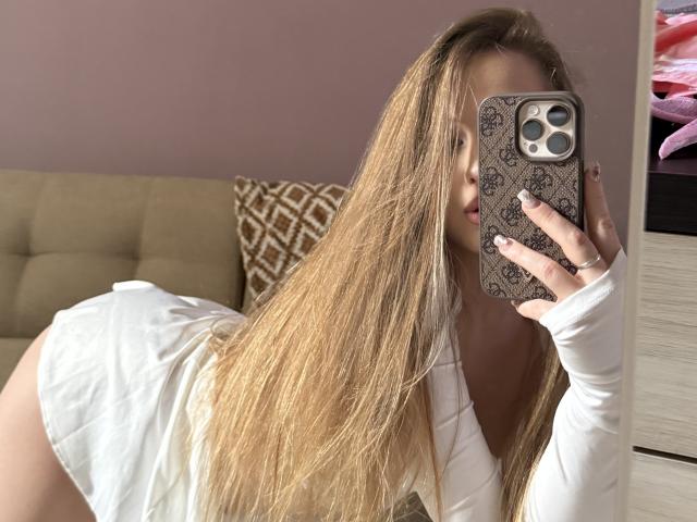 JoelleCanal with long blonde hair, white dress, and phone in mirror selfie, showing her ass and holding her phone with patterned case. Her face is partially visible.