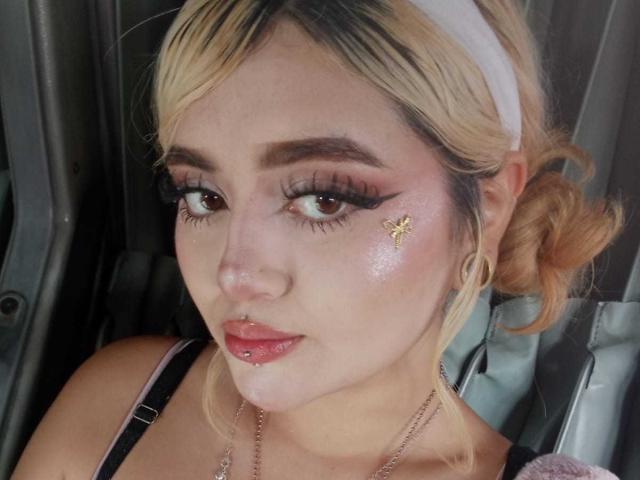 LyaMoon has blonde hair, a white headband, and a small butterfly sticker on her cheek. She's wearing long eyelashes and pink lipstick, with a slight sheen on her face.
