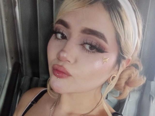 LyaMoon hat blondes Haar, dicke Augenbrauen und ein Nasenpiercing in Form eines Schmetterlings. Sie trägt eine goldene Kette, Ohrringe im Creolen-Stil und hat glänzende, rosafarbene Lippen. Ihr Make-up umfasst lange, dichte Wimpern..