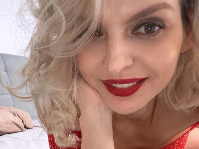 Close-up of DouceAmelia with wavy blonde hair and bright red lipstick. She's winking with her right eye and resting her chin on her right hand. Her red sleeve has white polka dots. She has fair skin, dark eyebrows, and subtle makeup. Her right hand partially covers her face. The background is a blurry, light-colored bedroom. The image has a casual, intimate feel.