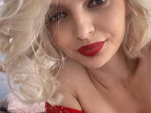 Close-up of DouceAmelia with curly blonde hair, fair skin, and bright blue eyes. She has bold red lipstick and a slight smile. She's wearing a red, sparkly, off-shoulder top. Her hair is slightly messy, and the light source is soft, creating gentle shadows. The focus is on her face and upper chest. The image is crisp with a slight blur in the background. The composition emphasizes her facial features and red lipstick.