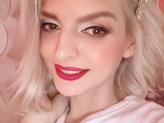 Close-up of DouceAmelia with platinum blonde hair, wearing bright red lipstick, and a pearl hairpin. Her light brown eyes are slightly almond-shaped with subtle eyeliner. She has a fair complexion with soft, natural-looking makeup. She's smiling slightly, showing a hint of teeth. She's wearing a white top. The image has a pinkish tone, and the focus is on her face. The hair is wavy and falls just past her shoulders.