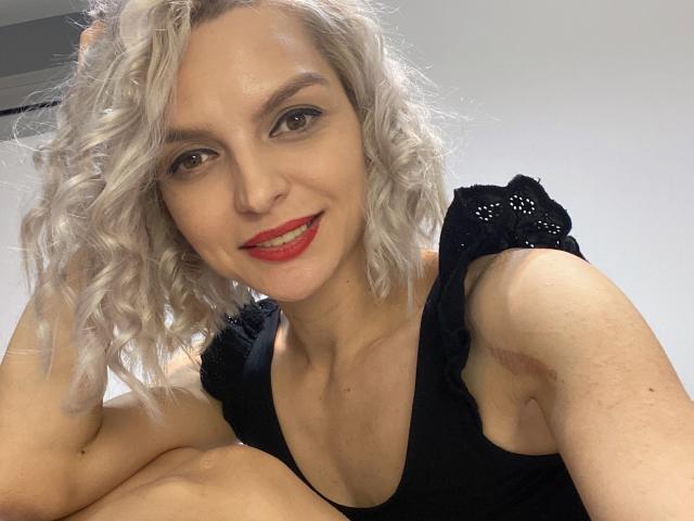 Close-up photo of DouceAmelia with wavy, platinum blonde hair, fair skin, and red lipstick. She's smiling, showing white teeth, with subtle eye makeup. She's wearing a black, sleeveless top with small, decorative holes. Her right arm is raised, hand in her hair. The left arm is partially visible with a small bruise on the upper arm. The background is plain and light-colored. Her facial expression is confident and friendly. The image has a casual, selfie-style composition.
