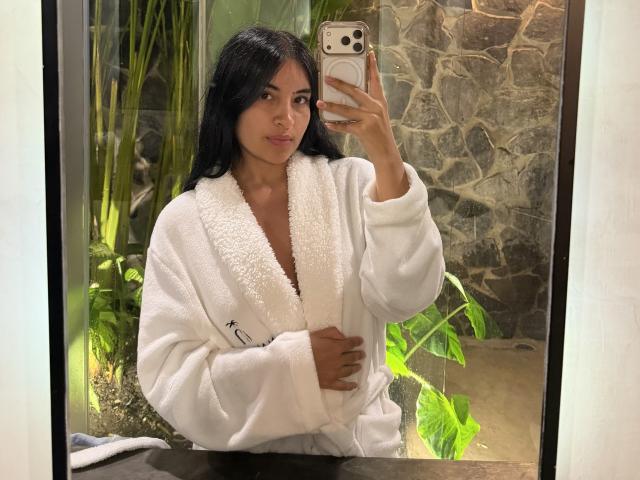 EmilyJensenn takes a mirror selfie with a smartphone. She has long, straight black hair and a light brown skin tone. She wears a fluffy white bathrobe with a small label on the left chest. Her right hand holds the phone, while her left hand rests on her chest. She has a neutral expression. The background includes green plants and a stone wall. The photo has a casual, modern style with a focus on her relaxed, everyday appearance.