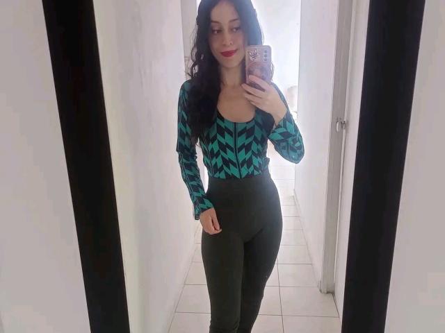 KendraSimons stands in a mirror selfie. She has long, wavy black hair and wears a green and black geometric patterned top with a deep V-neck, and dark, form-fitting pants. Her red lipstick contrasts with her fair skin. She holds a pink phone with a decorative case in her right hand. The mirror is framed in black, and the room has white tiled floors and white walls. She has a confident expression.