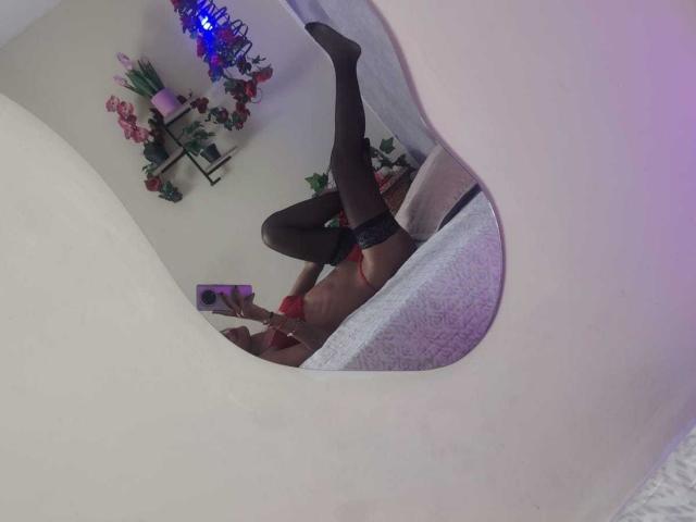 Mirrored selfie of ValerieWoods, lying on a bed with one leg raised. She wears black stockings and red lace panties. ValerieWoods holds a phone in her right hand, taking the photo. The mirror's white, curved frame is prominent. Background includes a shelf with flowers, pink candle, and blue LED lights. The bed has a white, textured blanket. ValerieWoods' skin is light brown, and her hair is dark. The photo has a casual, intimate vibe.
