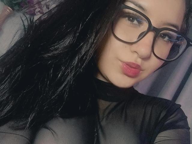 SusanMonroy has long black hair, wears black-framed glasses, and has pink lipstick. She's in a close-up selfie with a black mesh top. Her skin is light, and she looks directly at the camera with a slight smile. The image is sharp with a soft focus on her face, highlighting her glasses and lips. The texture of the mesh top contrasts with her smooth skin. The overall composition is centered on her face and upper torso.