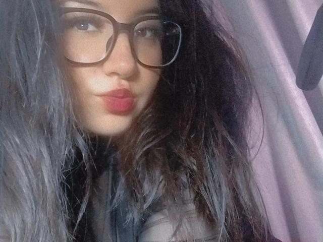 SusanMonroy with black hair, glasses, and pink lipstick, wearing a sheer black top, showing her tits slightly through the fabric. Her lips are puckered in a playful expression.