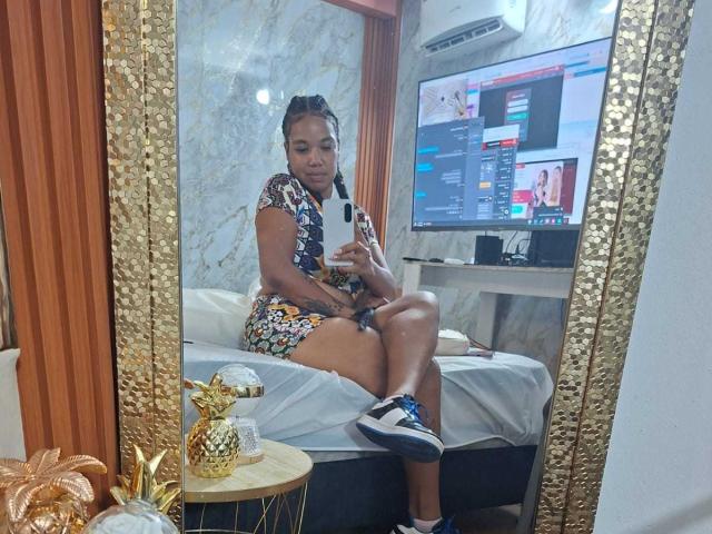 NaturaleBonyy with tan skin and braids, wearing a floral strapless top and white lace panties, taking a mirror selfie, showing her tits and pussy, holding a white phone.